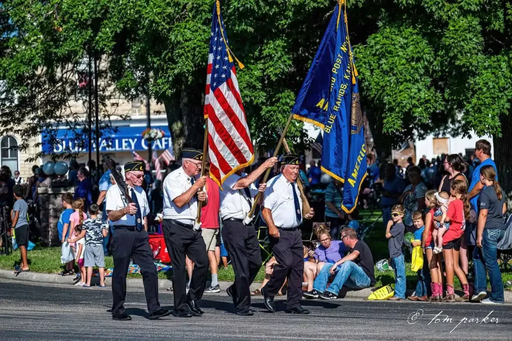 Veterans carrying the american flag and military flags in a parade, with spectators watching from the sidelines.