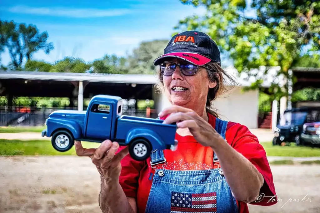 A woman wearing a cap and overalls holds up a blue toy truck outdoors.