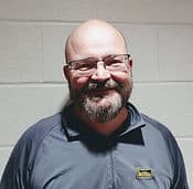 A smiling bearded man, identified as Jeff Keating, wearing glasses and a black polo shirt stands against a gray cinder block wall. He has a nametag on his shirt.