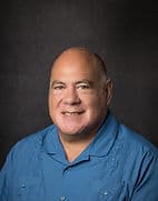 Portrait of a smiling man named Jerry Zayas with a bald head, wearing a blue collared shirt against a dark, textured background.