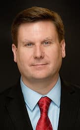 Portrait of a man named John Smith with short hair, wearing a dark suit, light blue shirt, and red tie, against a dark background. He is smiling slightly at the camera.