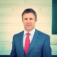 A man named Rob Peschel, dressed in a dark suit and red tie, smiling in front of a light brick wall.