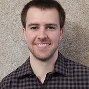 A young man with a friendly smile, wearing a plaid shirt, stands without additional details like an associated industry against a beige textured background.